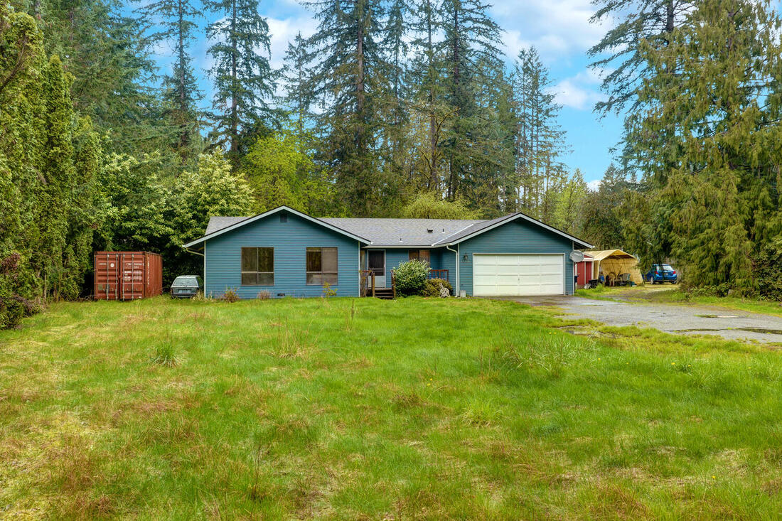 Huge front yard and blue 1-story home with treed background