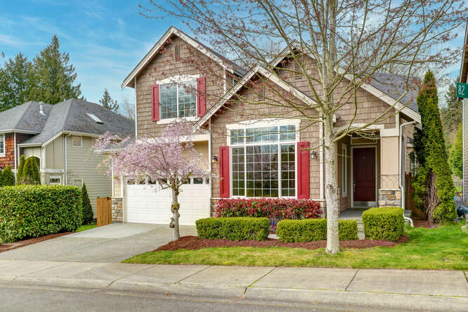 Front of the house with beige siding color with red shutters by big windows. Cherry blossom tree is blooming with pink flowers.